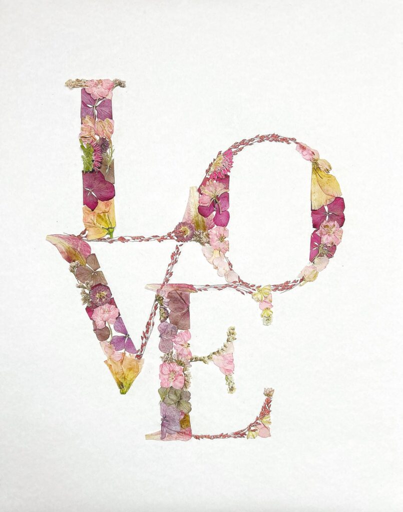 Das Wort "LOVE" in Großbuchstaben, gebildet aus getrockneten Blumen und Blüten in rosa, violett und gelben Farbtönen auf weißem Hintergrund.
