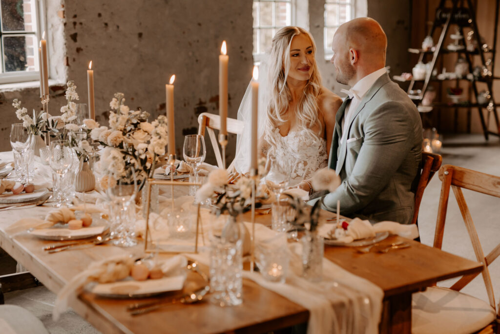 Brautpaar sitzt an einem festlich gedeckten Holztisch mit Kerzen und Blumen, die Braut trägt ein weißes Kleid mit Spitzendetails, der Bräutigam einen grauen Anzug, beide blicken sich liebevoll an.