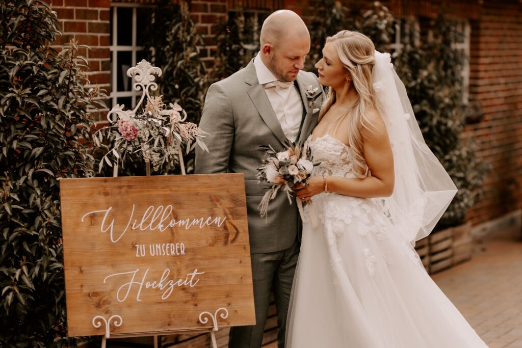 Ein Brautpaar in Hochzeitskleidung steht nah beieinander vor einem Schild mit der Aufschrift "Willkommen zu unserer Hochzeit" an einer mit Efeu bewachsenen Backsteinwand.