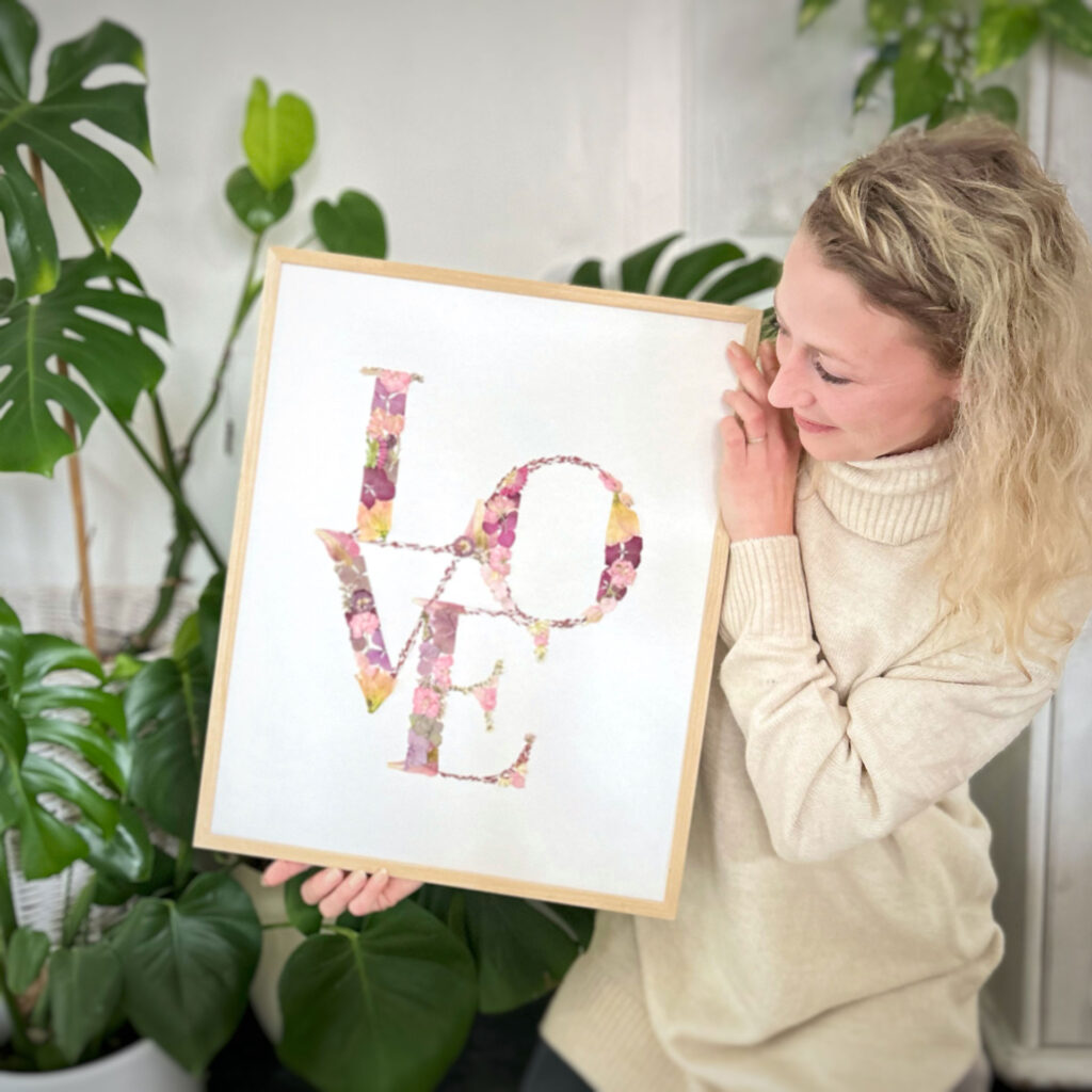 Frau mit hellem Pullover hält und betrachtet einen Bilderrahmen mit dem Schriftzug LOVE, der aus bunten Blumen besteht, vor einem Hintergrund mit grünen Zimmerpflanzen.